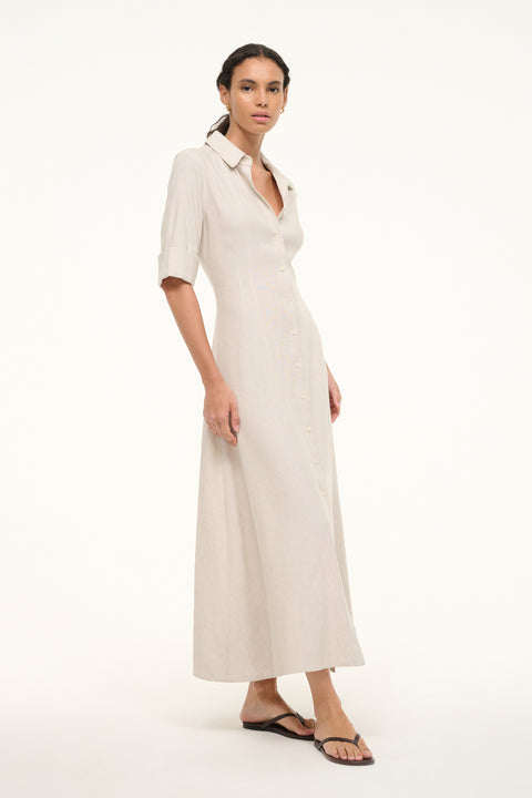 Side view of a woman wearing a cream linen maxi dress that features half sleeves, a collar, and button down detail down he front, paired with sandals.  