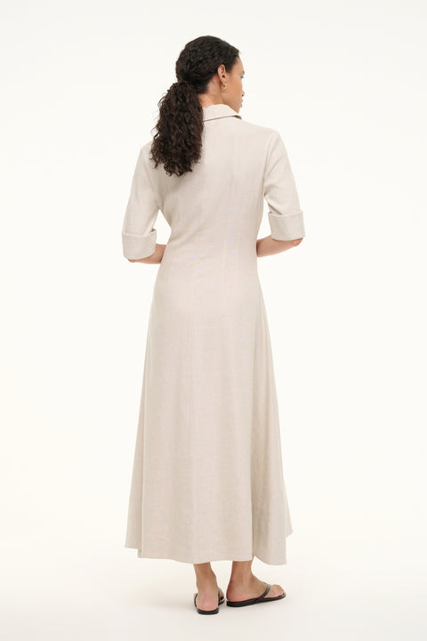 Back view of a woman wearing a cream linen maxi dress that features cuffed half sleeves and a collar, paired with sandals. 
