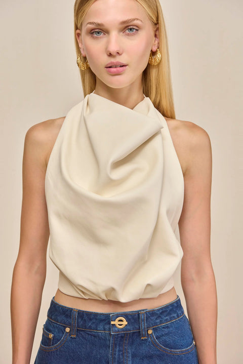 Close up of a woman wearing a cream, faux leather halter top that drapes throughout the bodice, paired with blue jeans. 