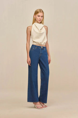Woman wearing a cream, faux leather halter top that drapes throughout the bodice, paired with blue jeans. 
