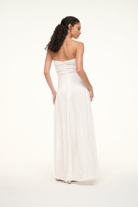 Back view of a woman wearing a white strapless maxi dress that features a ruched bodice, a curved yoke at the hip, and a full skirt. 