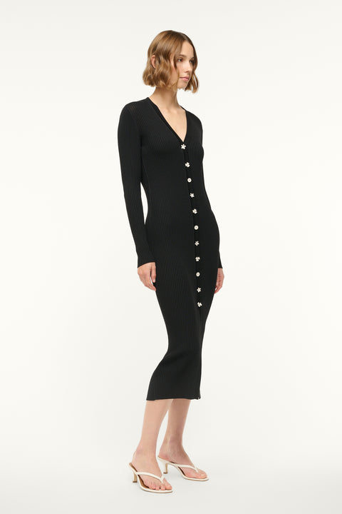 Side view of a woman wearing a black ribbed-knit, long sleeve midi dress that features silver toned floral buttons down the front, paired with white sandals. 