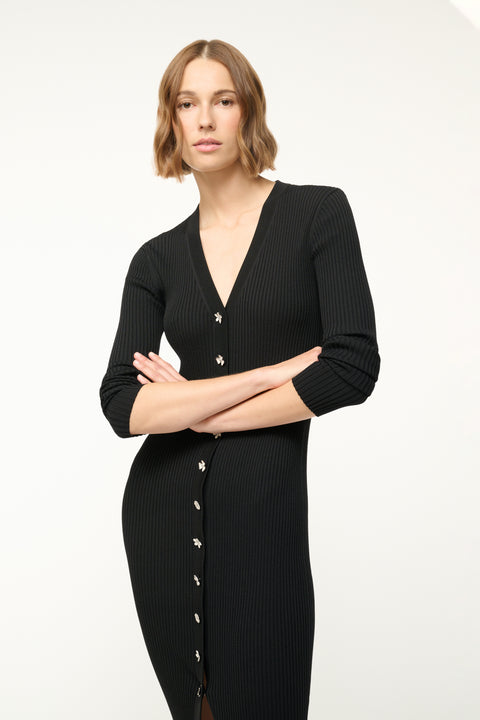 Close up of a woman wearing a black ribbed-knit, long sleeve midi dress that features silver toned floral buttons down the front, standing with her arms crossed over her chest.