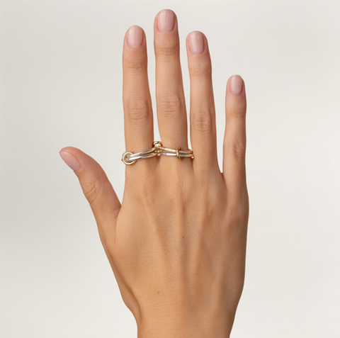 A woman's hand wearing a Spinelli Kilcollin ring that features a slim sterling silver band, a central 18k yellow gold band with channel set white diamonds, another slim sterling silver band, and a thicker sterling silver band, all connected by three 18k yellow gold connector rings., with two of the rings on the index finger and two of the rings on the middle finger. 