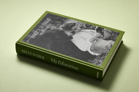 Coffee table book title "Bruce Weber: My Education" that depicts a black and white picture of a man sitting next to a bear with a green border and green spine. 