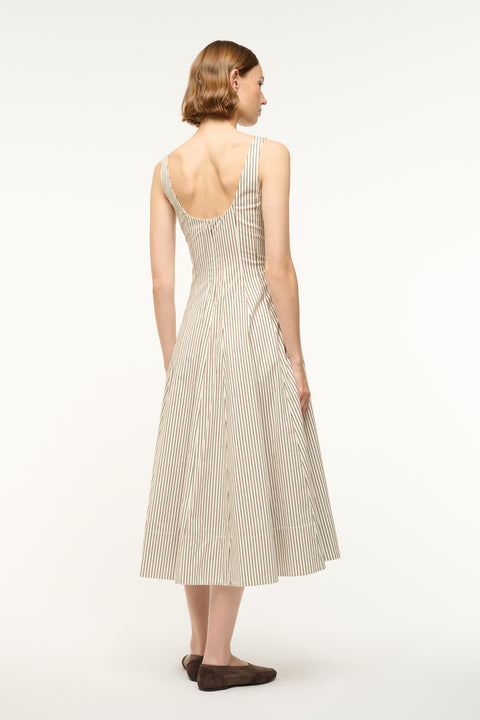 Back view of a Woman wearing a midi dress that boasts a scoop back, corset-style bodice, full, and full A-line skirt in a cream and brown micro stripe pattern, paired with flats. 