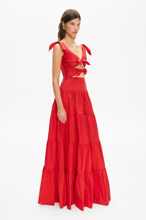 Side angle of a woman wearing a red v-neck maxi dress that features ties at the shoulders and two cutouts on the torso with a knot detail. 