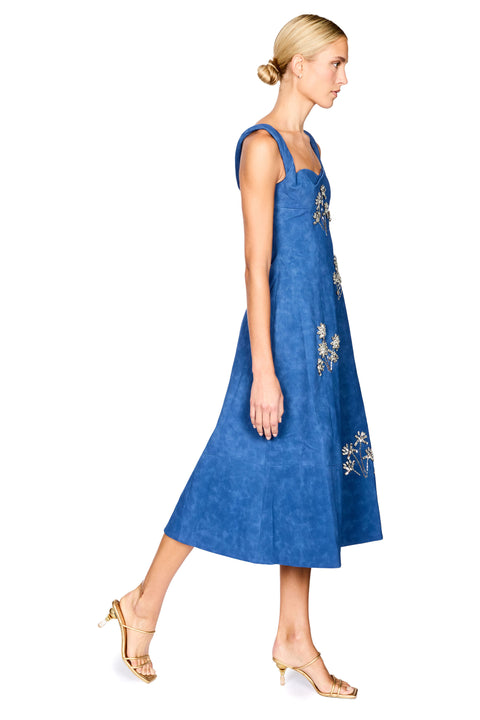 A woman modeling a sleeveless blue midi dress with silver floral embroidery, shown in profile against a plain white background.