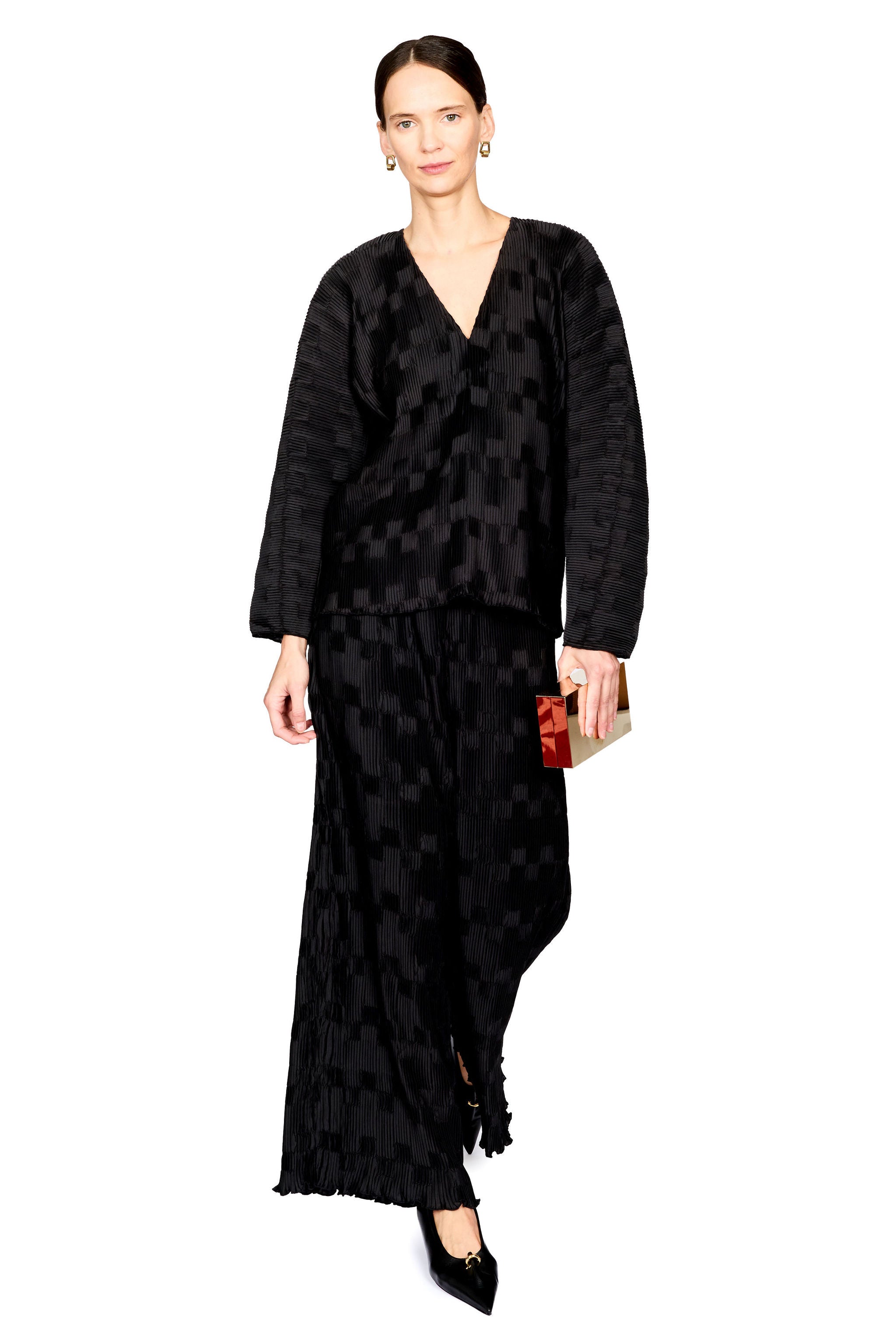 A model wearing a black long-sleeve maxi dress with a textured geometric pattern and fringe hem, holding a small clutch against a solid white background.