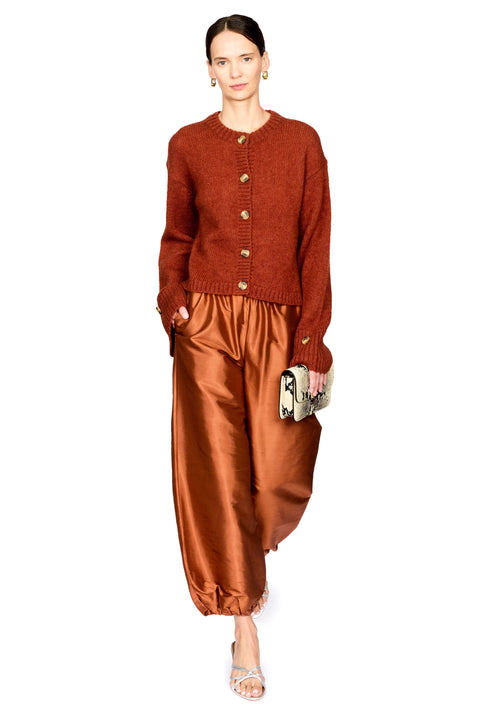 A model is shown from the head to toe wearing a rust-orange knit button-front cardigan, matching rust-orange satin pants, and holding a small metallic clutch against a plain white background.