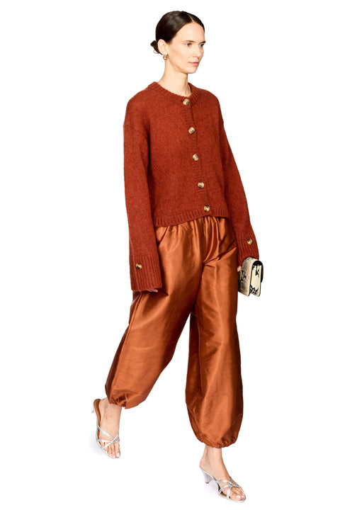 A model wearing a rust-orange knit cardigan and matching satin trousers, styled with silver sandals and holding a small patterned clutch against a plain white background.