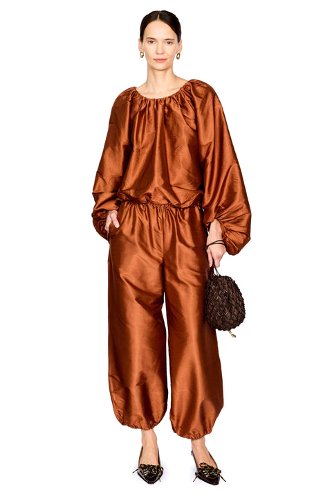 A model wearing a shiny rust-brown satin two-piece set consisting of a loose long-sleeve top and matching wide-leg pants, holding a small dark textured handbag against a plain white background.