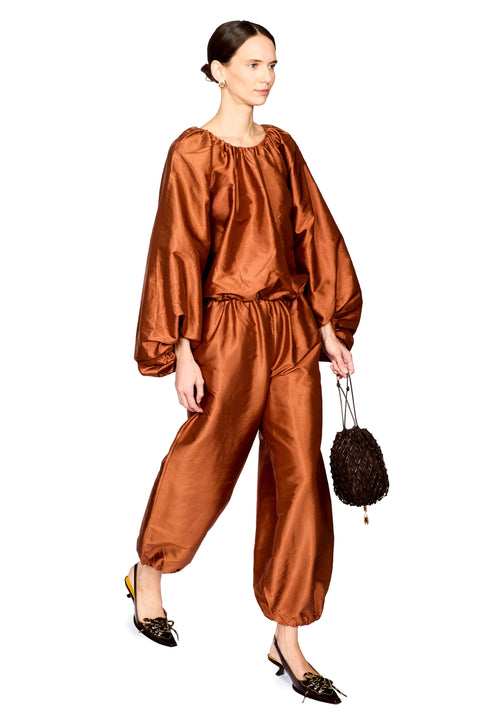 A model wearing a shiny rust-brown satin two-piece set with a loose, voluminous top and matching baggy pants, holding a small dark brown textured handbag against a plain white background.