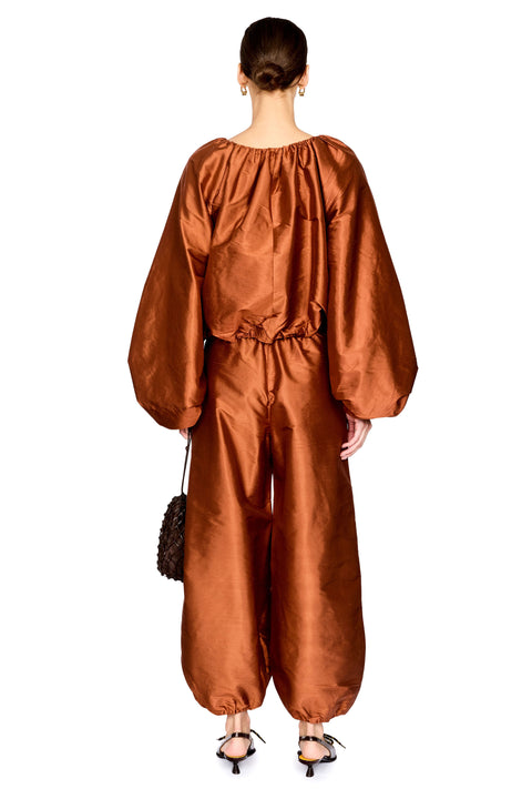 A model stands against a white background wearing a metallic copper-brown satin two-piece outfit with voluminous balloon sleeves and gathered harem-style pants, holding a small dark woven handbag.
