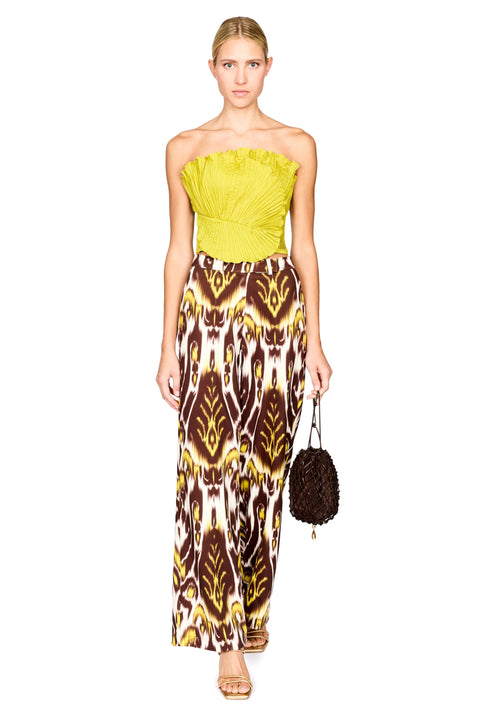 A model stands against a white background wearing a strapless bright lime top and wide-leg trousers with a brown, yellow, and white ikat pattern. She is holding a small dark brown textured handbag.