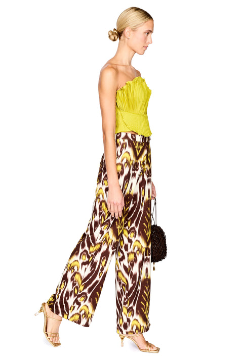 A model stands against a white background wearing a strapless bright lime top and wide-leg trousers with a brown, yellow, and white ikat pattern. She is holding a small dark brown textured handbag.