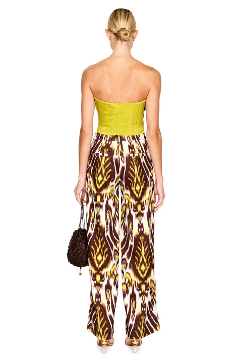 A model stands against a white background wearing a strapless bright lime top and wide-leg trousers with a brown, yellow, and white ikat pattern. She is holding a small dark brown textured handbag.