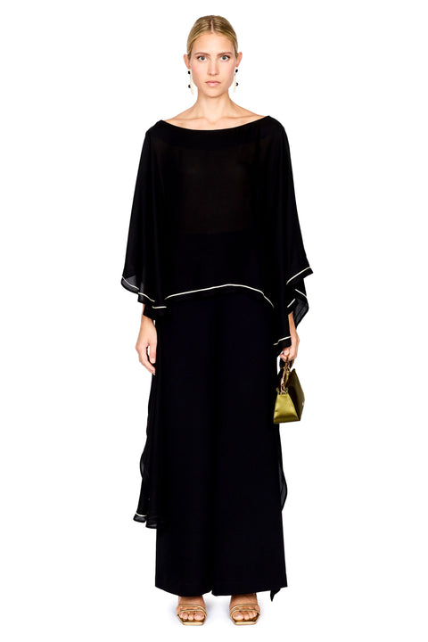 A woman modeling a black pants and a matching top with wide draped sleeves and thin white piping accents, holding a small green handbag against a plain white background.