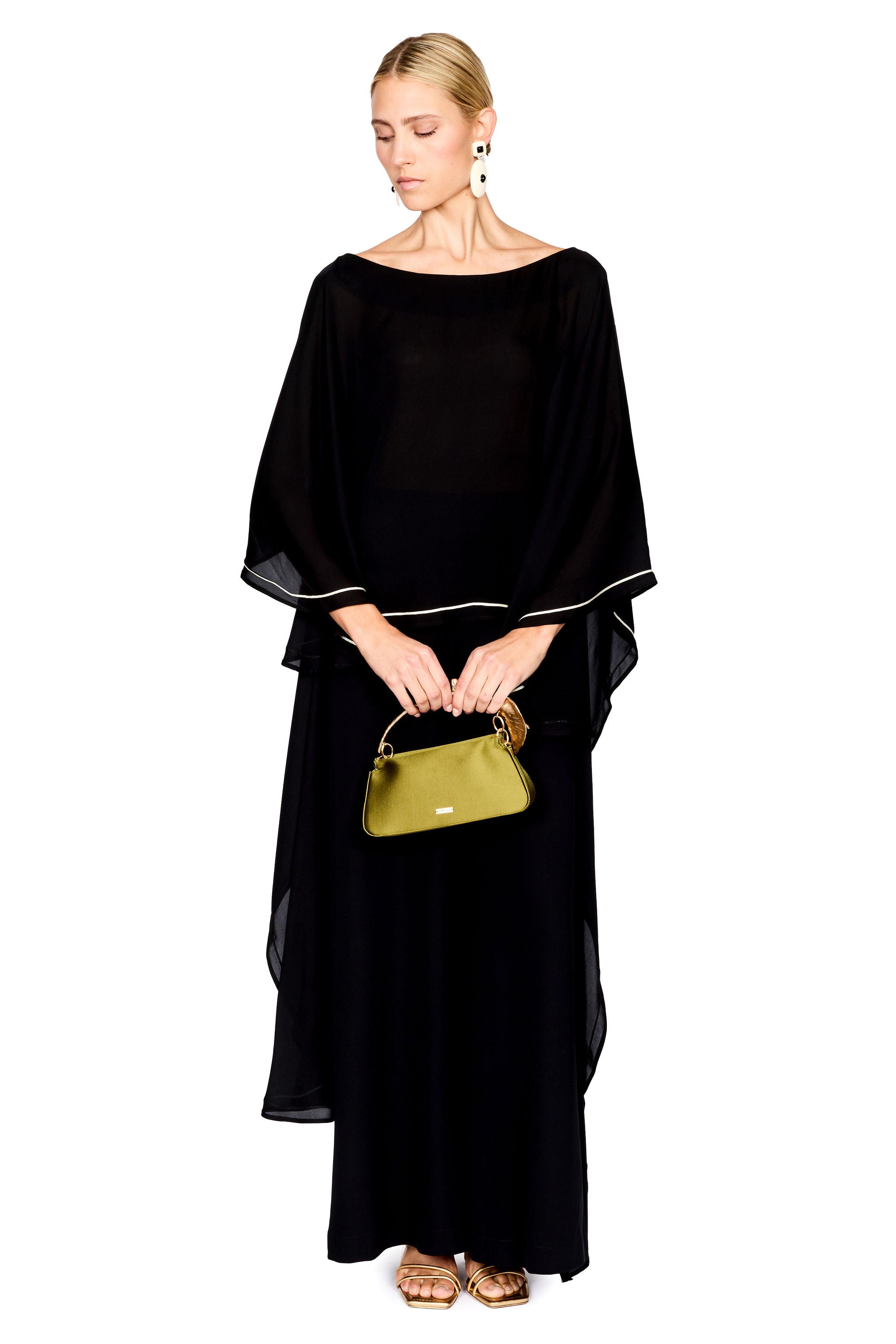 A woman modeling a black pants and a matching top with wide draped sleeves and thin white piping accents, holding a small green handbag against a plain white background.