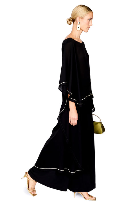 A woman modeling a black pants and a matching top with wide draped sleeves and thin white piping accents, holding a small green handbag against a plain white background.