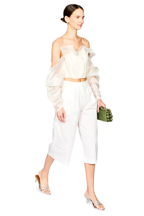A model wearing a white two-piece outfit consisting of an off-the-shoulder sheer long-sleeve crop top and matching white cropped trousers, holding a small green textured clutch.