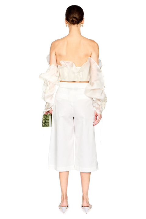A model shown from the back wearing a white off-the-shoulder crop top with dramatic puffed sleeves and matching white wide-leg cropped pants, holding a small green patterned clutch against a plain white background.