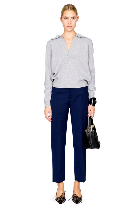 A model stands against a white background wearing a light gray wrap-style knit sweater, navy blue cropped trousers, black pointed flats, and holding a black handbag.