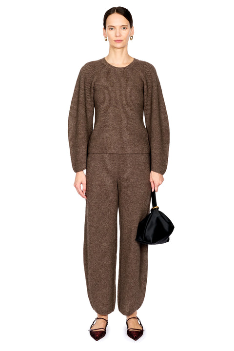 A model stands against a plain white background wearing a matching brown knit long-sleeve top and wide-leg trousers set, holding a small black handbag.