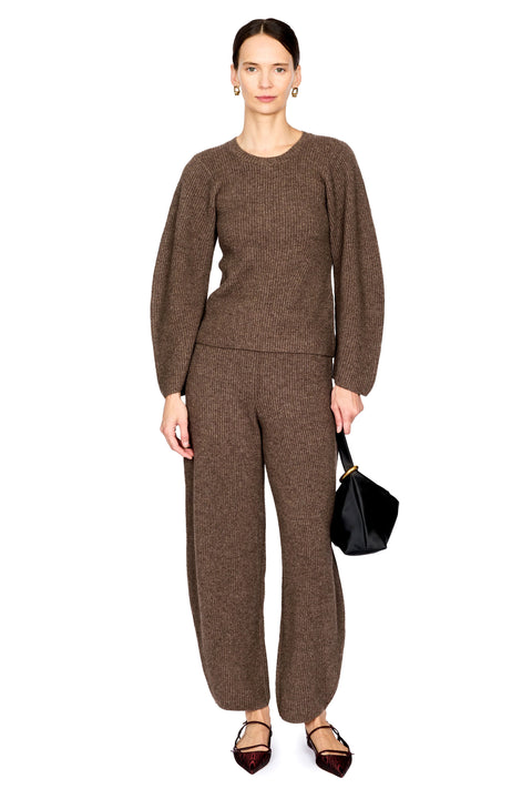 A model stands against a white studio background wearing a matching brown knit two-piece set consisting of a long-sleeve crewneck sweater and wide-leg trousers. She is holding a small black handbag and wearing dark pointed flats.