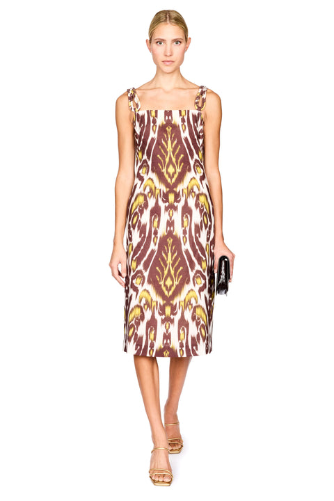 A woman modeling a midi dress with a brown, gold, and cream ikat pattern, paired with gold sandals and holding a small black clutch, set against a plain white background.