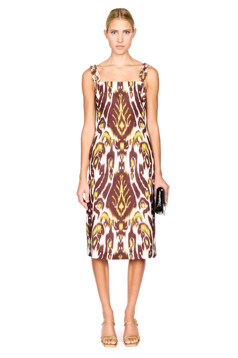 A woman modeling a midi dress with a brown, gold, and cream ikat pattern, paired with gold sandals and holding a small black clutch, set against a plain white background.