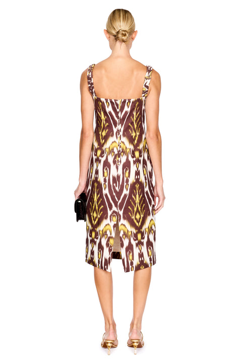 A woman modeling a midi dress with a brown, gold, and cream ikat pattern, paired with gold sandals and holding a small black clutch, set against a plain white background.