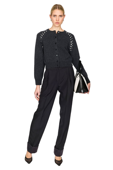 A model stands against a white studio background wearing a dark charcoal gray knit cardigan with silver stud embellishments on the shoulders, paired with black trousers and black pointed shoes, while holding a black and white handbag.