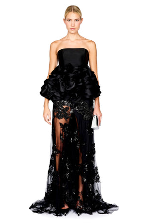 A model wearing a strapless top with a dramatic ruffled bodice, a long black skirt with a sheer floral sequin embellished lace overlay, posed against a plain white studio background.