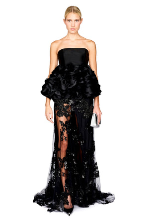 A model wearing a strapless top with a dramatic ruffled bodice, a long black skirt with a sheer floral sequin embellished lace overlay, posed against a plain white studio background.