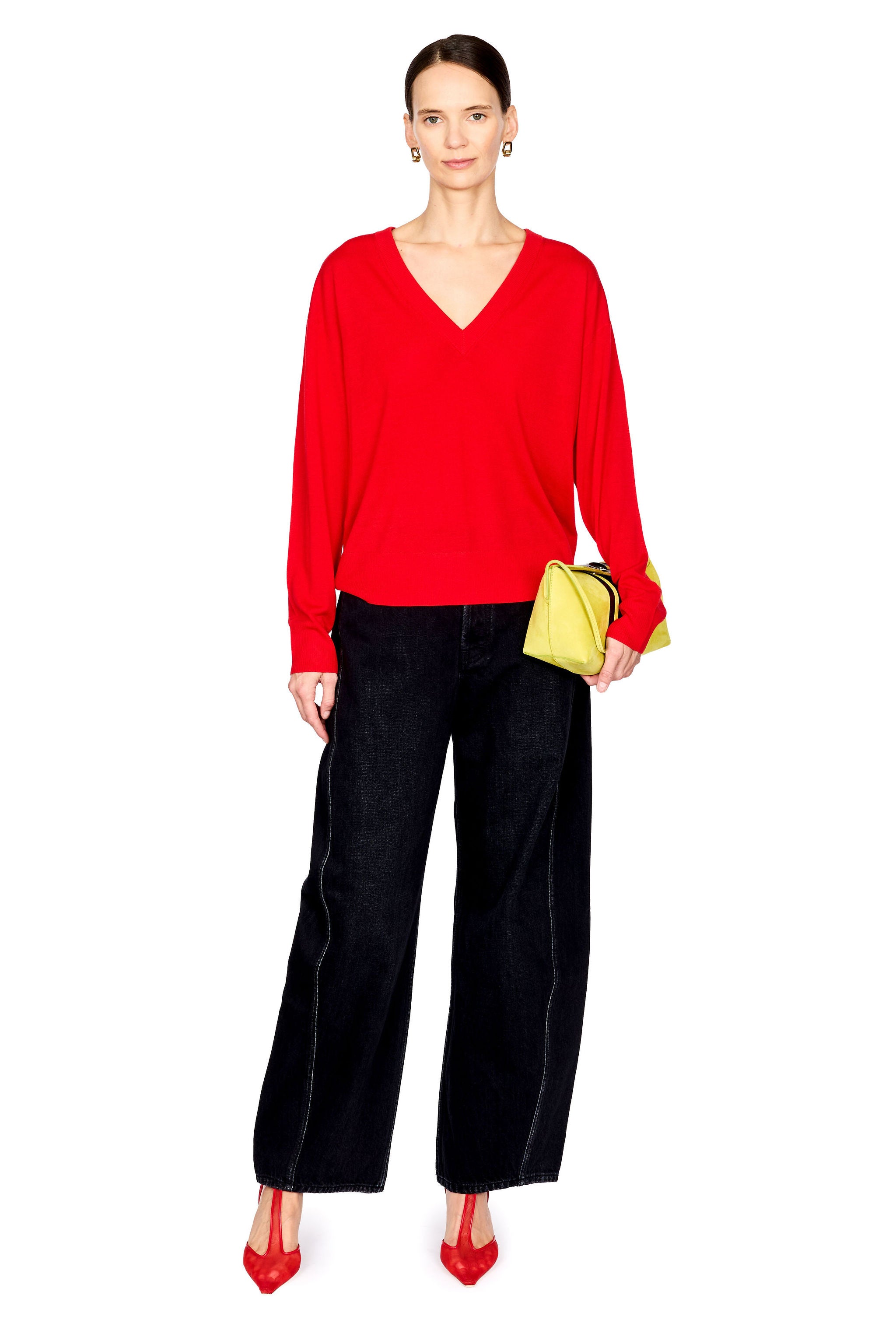 A model stands against a white background wearing a bright red long-sleeve V-neck sweater, black wide-leg trousers, red pointed heels, and holding a small yellow clutch.
