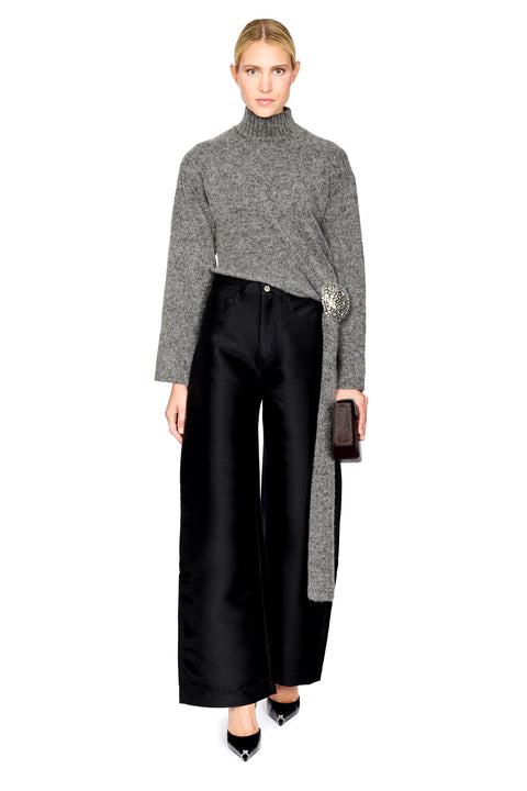 A model stands against a white studio background wearing a gray marled turtleneck sweater draped over black wide-leg satin trousers, with pointed black heels and a small dark clutch.