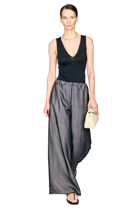 A model wearing a black sleeveless V-neck top and wide-leg gray pinstriped trousers, holding a small cream-colored handbag against a plain white background.