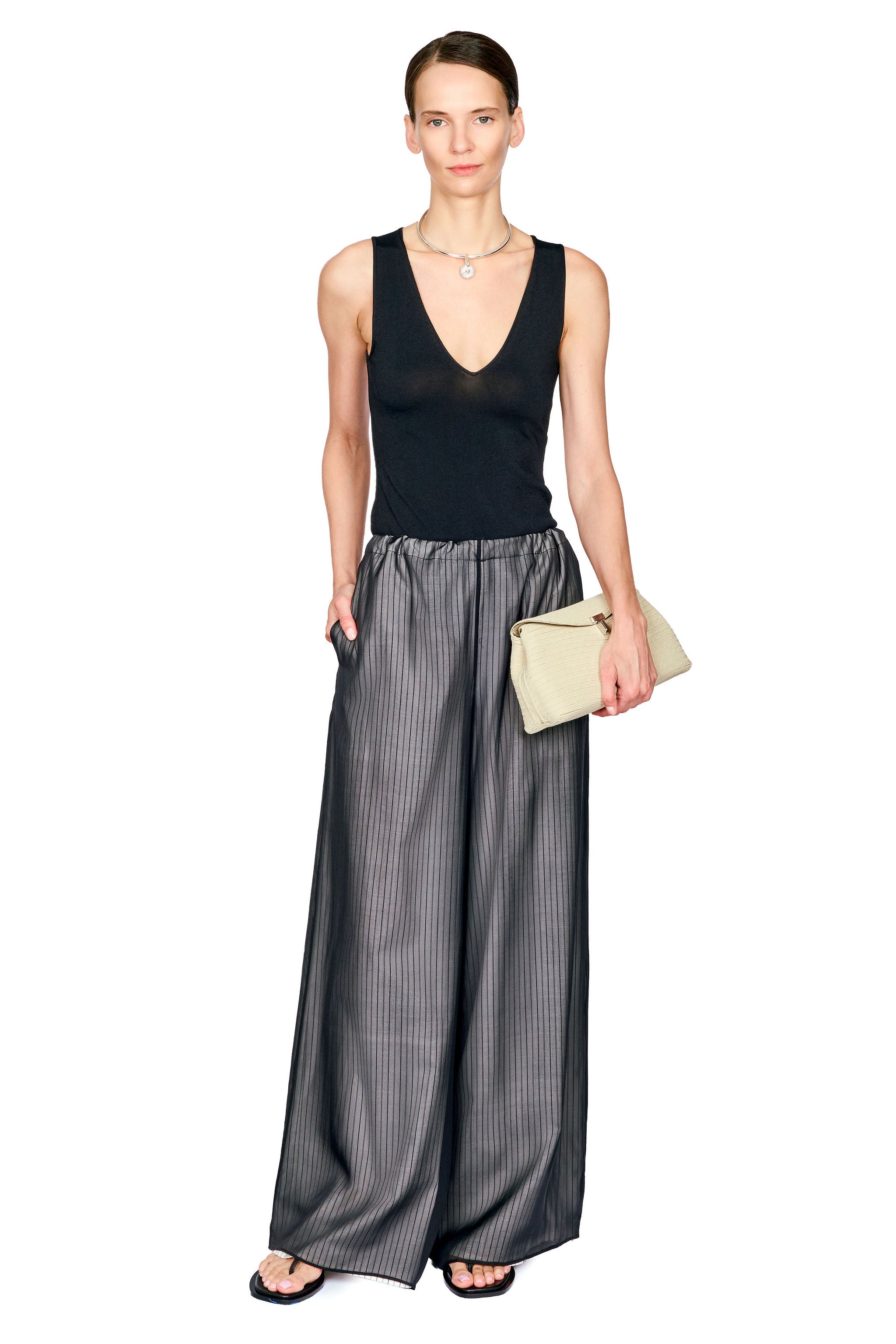A model stands against a white background wearing a black sleeveless V-neck top and wide-leg charcoal gray pinstriped trousers, holding a small beige clutch.