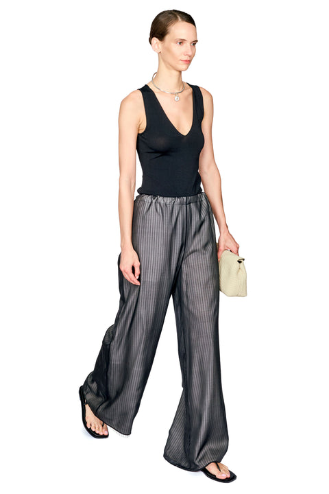 A woman modeling a sleeveless black V-neck top and wide-leg pinstriped trousers in a studio setting, holding a small beige clutch.