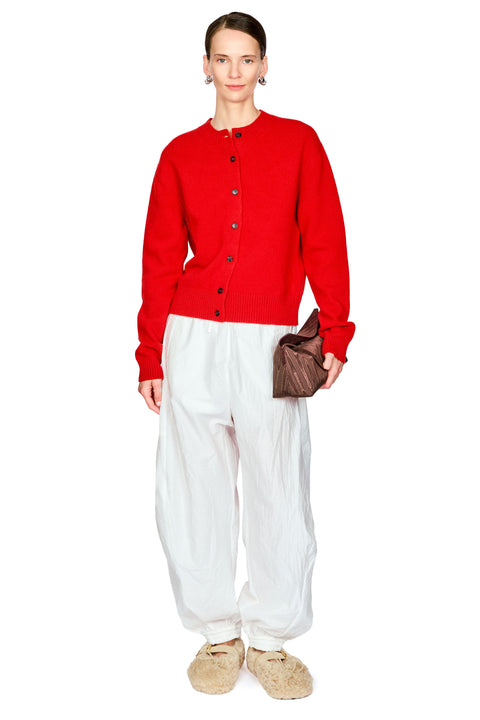 A model stands against a white background wearing a bright red button-front cardigan, loose white pants, beige slippers, and holding a small brown bag.