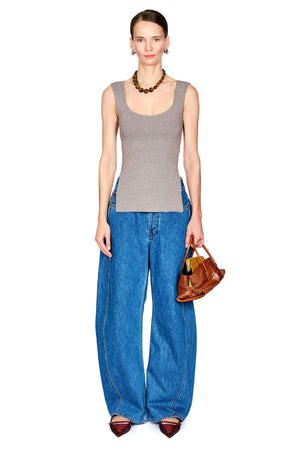A model stands against a white background wearing a gray ribbed tank top, wide-leg blue jeans, brown pointed shoes, a brown beaded necklace, and holding a brown leather handbag.