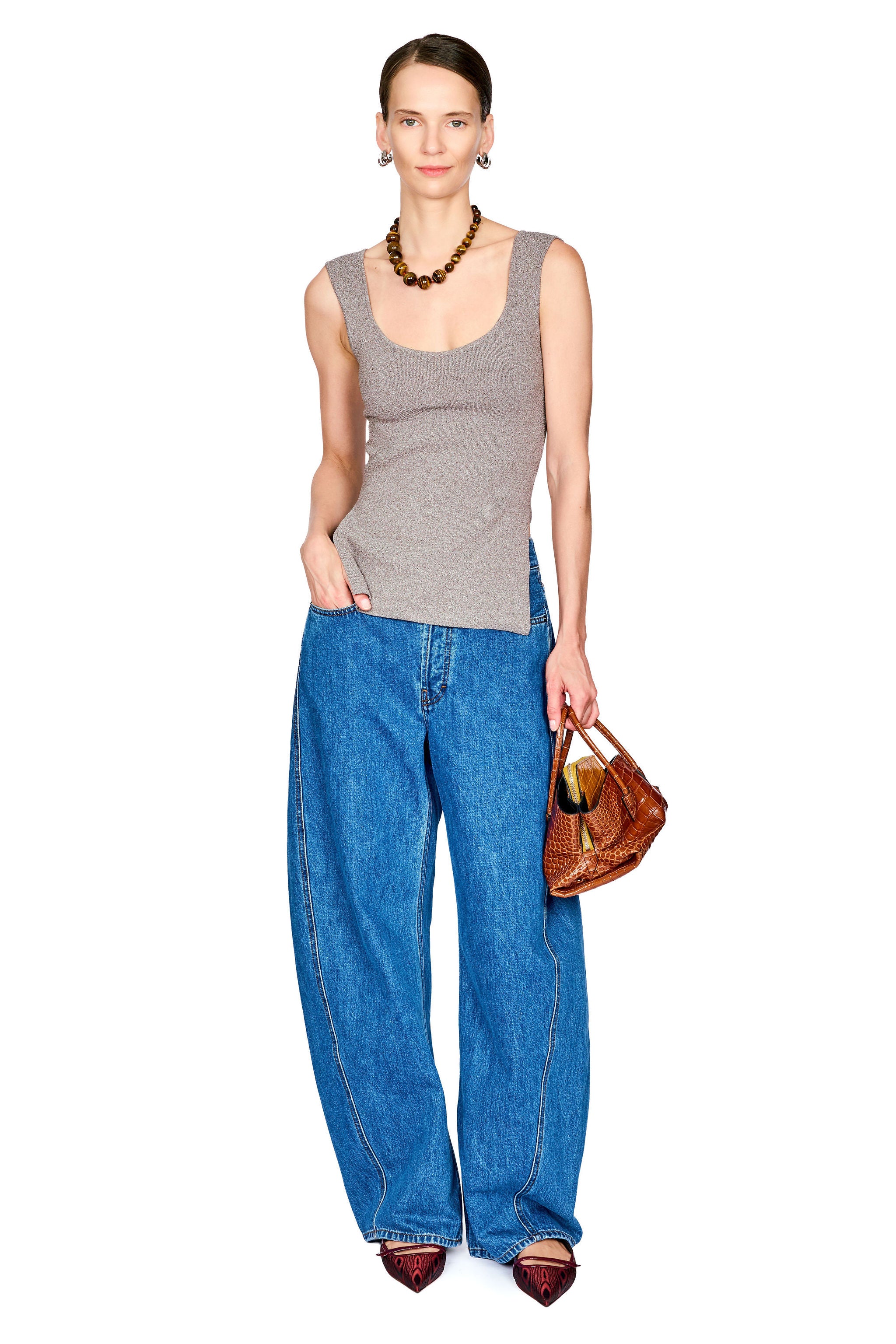 A woman modeling a gray ribbed tank top and wide-leg blue jeans, holding a brown leather handbag against a plain white background.