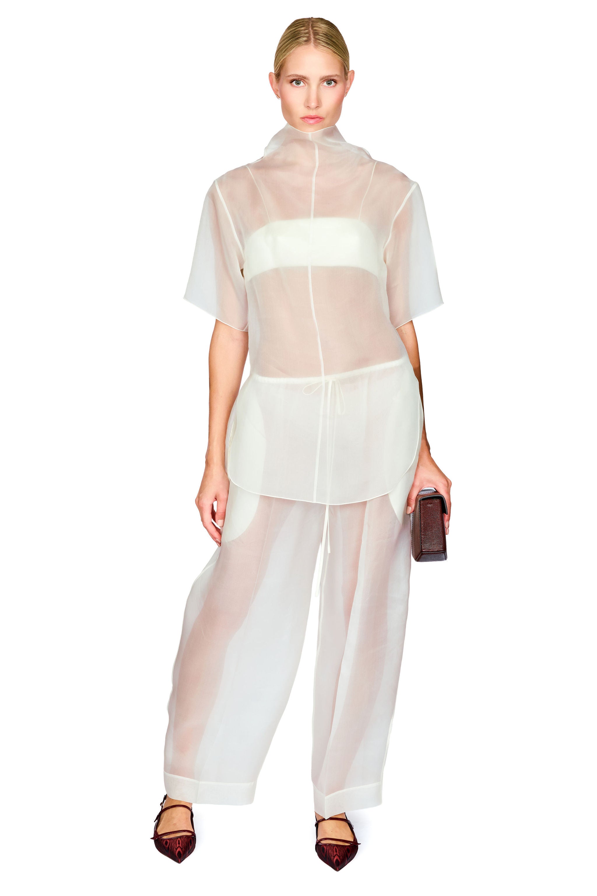 A model wearing a sheer white high-neck short-sleeve top and matching wide-leg sheer pants, holding a small dark clutch against a plain white background.