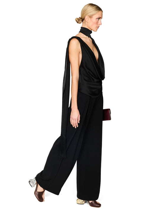 A woman modeling a black sleeveless draped jumpsuit with a high neck and wide-leg pants, shown in profile against a plain white background.