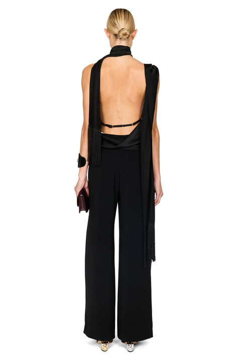 A model shown from the back wearing a black sleeveless wide-leg jumpsuit with a high neck and draped fabric detail, paired with black strappy heels, on a solid white background.