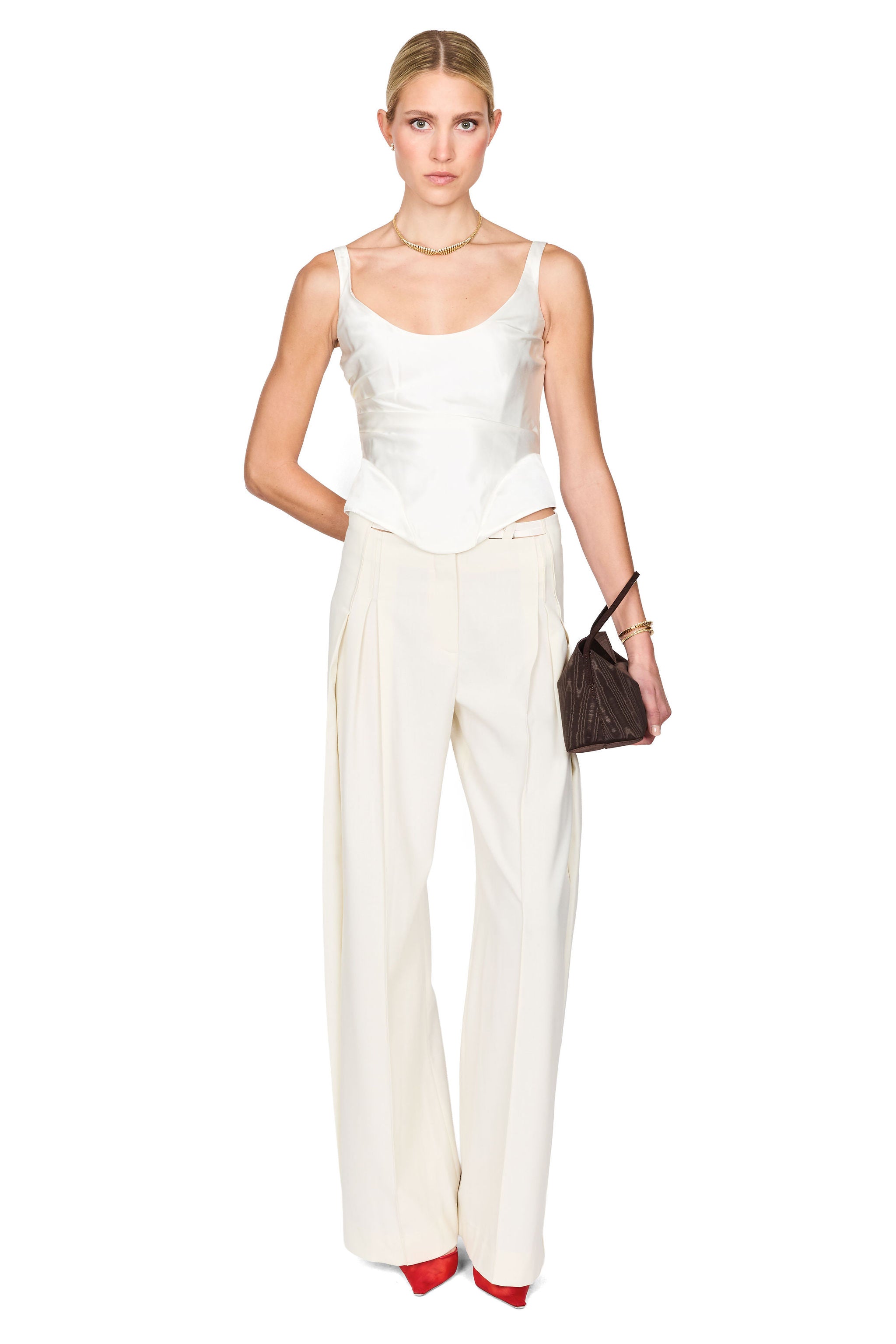 A model wearing a white sleeveless camisole top and matching white wide-leg trousers, holding a small dark brown handbag, against a solid white background.