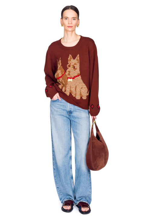Person wearing a brown sweater with dog designs, blue jeans, and carrying a brown bag on a white background with one hand in pocket.