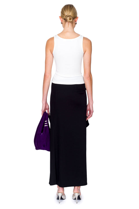 Woman wearing a white cutout top with a crystal pendant and black skirt with a purple bag on a white background from the back.