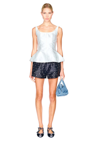 A model stands against a white background wearing a white sleeveless peplum top, black shorts with white polka dots, and black strappy sneaker flats, while holding a blue handbag.